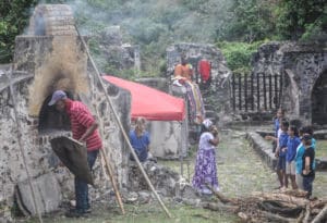 Donald Christopher cooks up dumb bread old-fashioned style, while in the background Allegra Christopher teaches bamboula dancing to Antilles School students and Mocko Jumbie Yisrael Petersen takes a rest on a ruin wall.