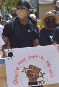 In Roosevelt Park, A protester holds up a sign referencing Queen Mary, one of the leaders of the Fireburn protest of 1878. (Source photo by Kyle Murphy)